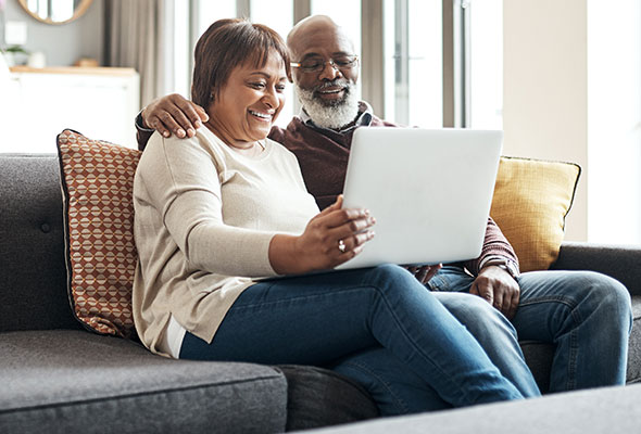 Man and woman sitting on a couch smiling while reviewing their preventative service plan on a laptop.