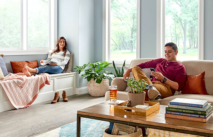 Woman reading on a window seat, man on a couch smiling at how much money their new furnace has saved them on his phone