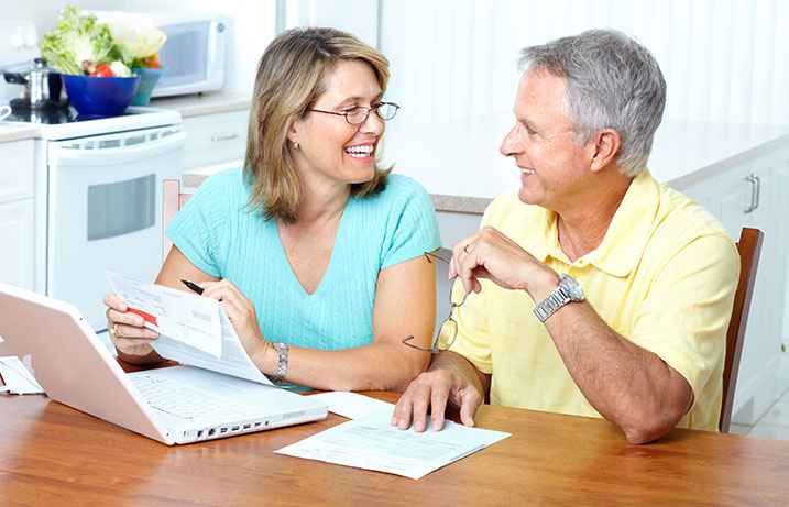 Man holding glasses and woman at a laptop sitting at the kitchen table smiling about the rebate check she is holding