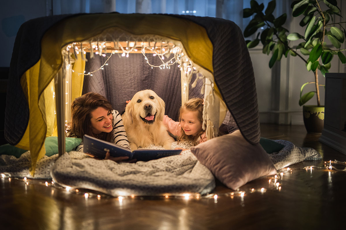 Woman, young girl and dog under a table draped with string lights and blankets reading, while their heat pump keeps them cozy