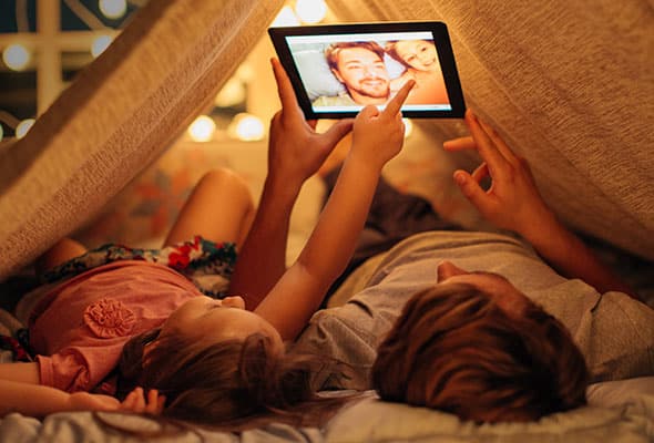 Two people looking at an iPad while laying under an indoor blanket tent, cozy, warm and safe during a power outage