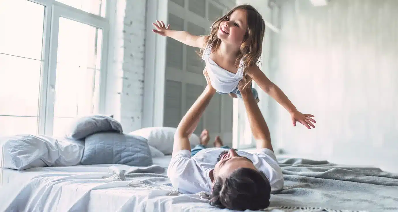 Man laying on bed holding child over him, flying her like an airplane enjoying their new central air conditioning from Central Cooling & Heating in Woburn, MA