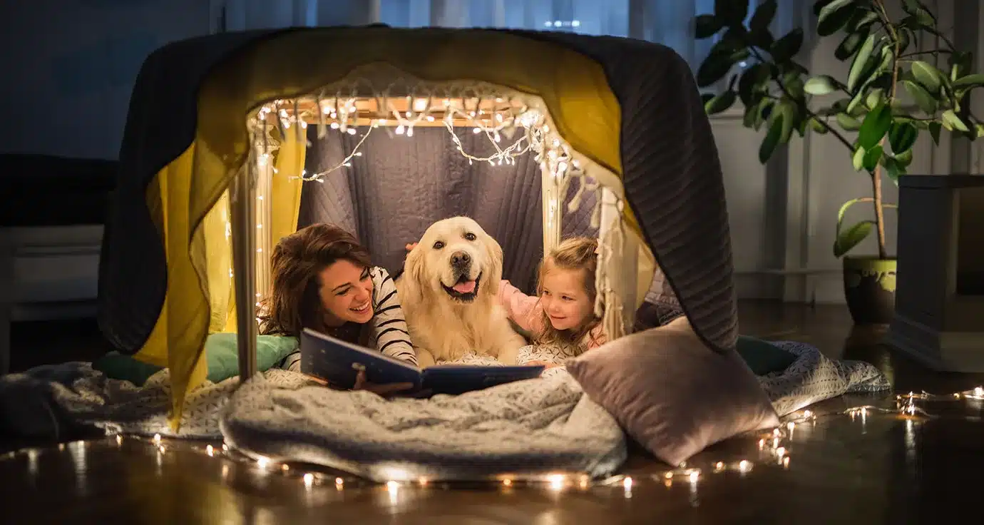 Woman, young girl and dog under a table draped with string lights and blankets reading, while their heat pump keeps them cozy