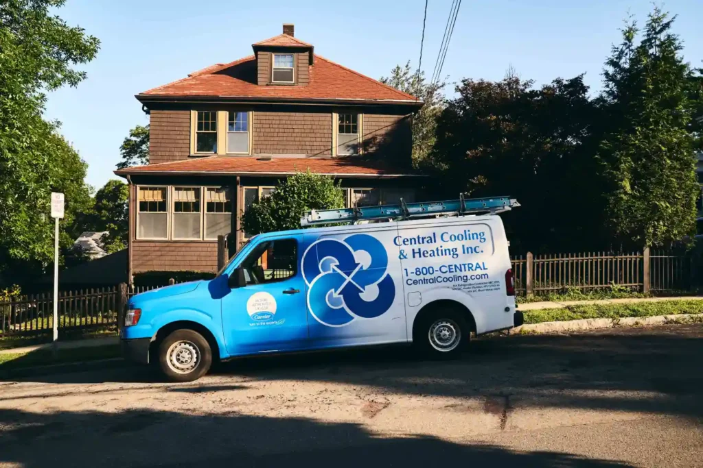 A blue Central Cooling & Heating van is parked on a residential street in front of a brown house, with a ladder on its roof.