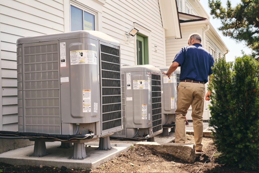 A technician inspects three outdoor Carrier air conditioner units installed beside a white building.