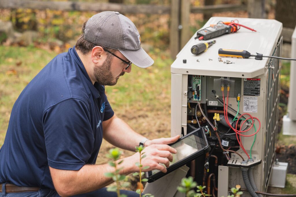 A technician wearing a cap and glasses inspects and repairs the wiring of an outdoor heat pump HVAC unit using tools and a tablet.