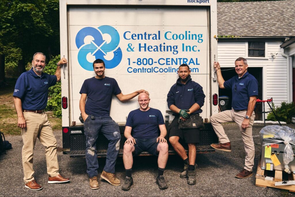 Five men wearing navy shirts and khaki or dark pants pose in front of a Central Cooling & Heating Inc. truck parked on a driveway, representing their plumbing services. Tools and equipment are visible on the right.