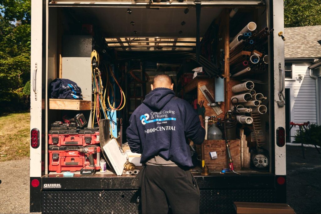 A person stands at the back of a work truck filled with tools, pipes, and equipment, wearing a hoodie with a plumbing and heating company logo.