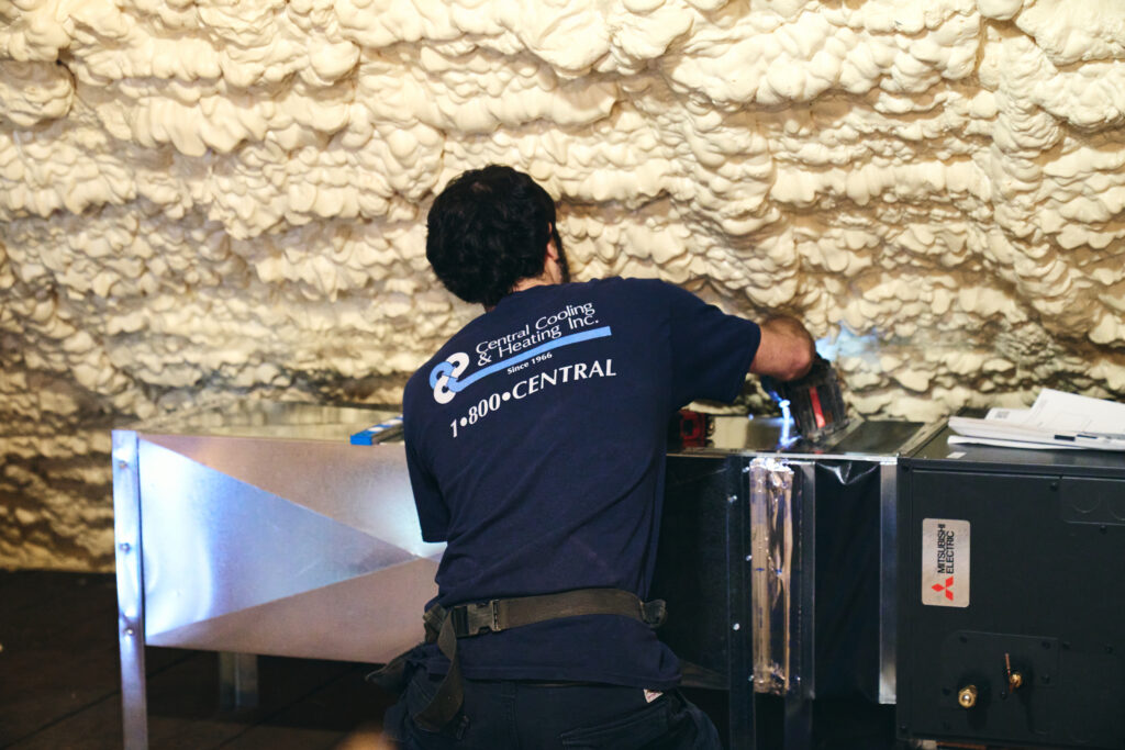Central Cooling & Heating technician installing heating system ductwork and air handler in attic with spray foam insulation