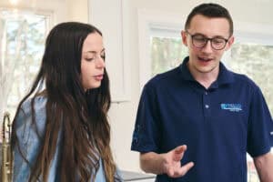 A man in a navy polo shirt speaks to a woman with long hair as they stand together indoors, near a kitchen sink and windows.