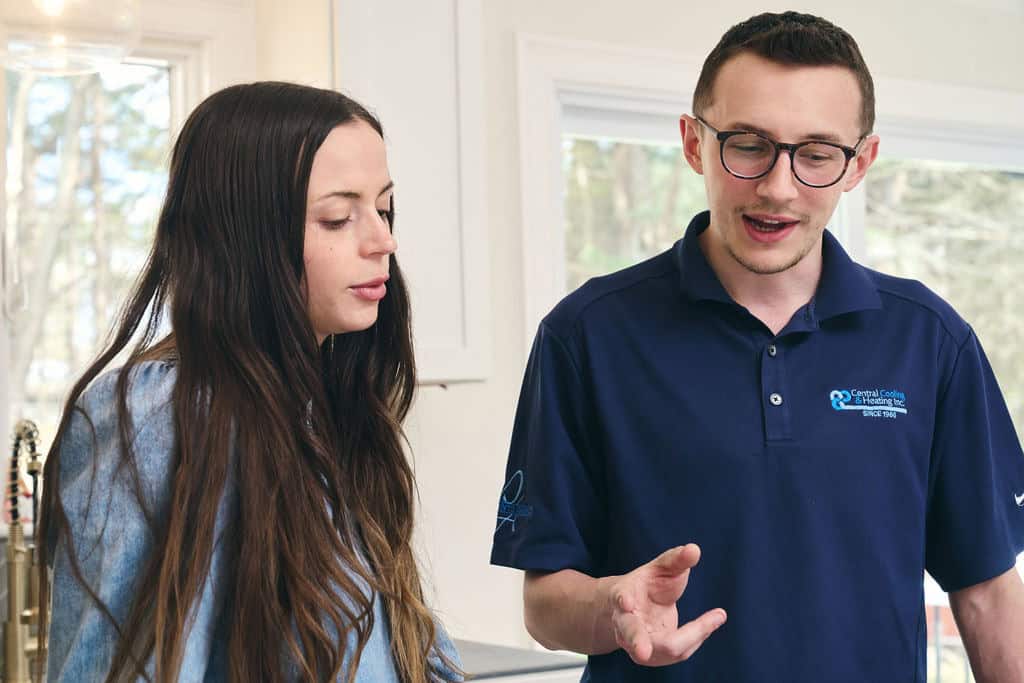 A man in a navy polo shirt speaks to a woman with long hair as they stand together indoors, near a kitchen sink and windows.