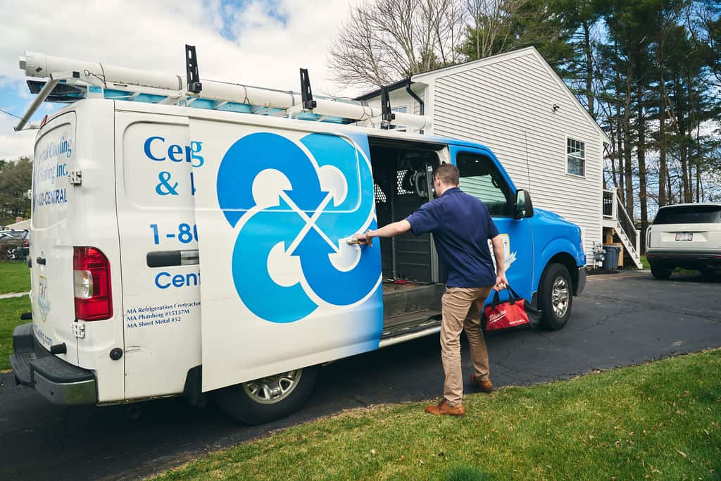 A man in a blue shirt opens the side door of a white service van parked in a driveway, holding a red tool bag—ready to provide HVAC and plumbing services in Woburn MA.
