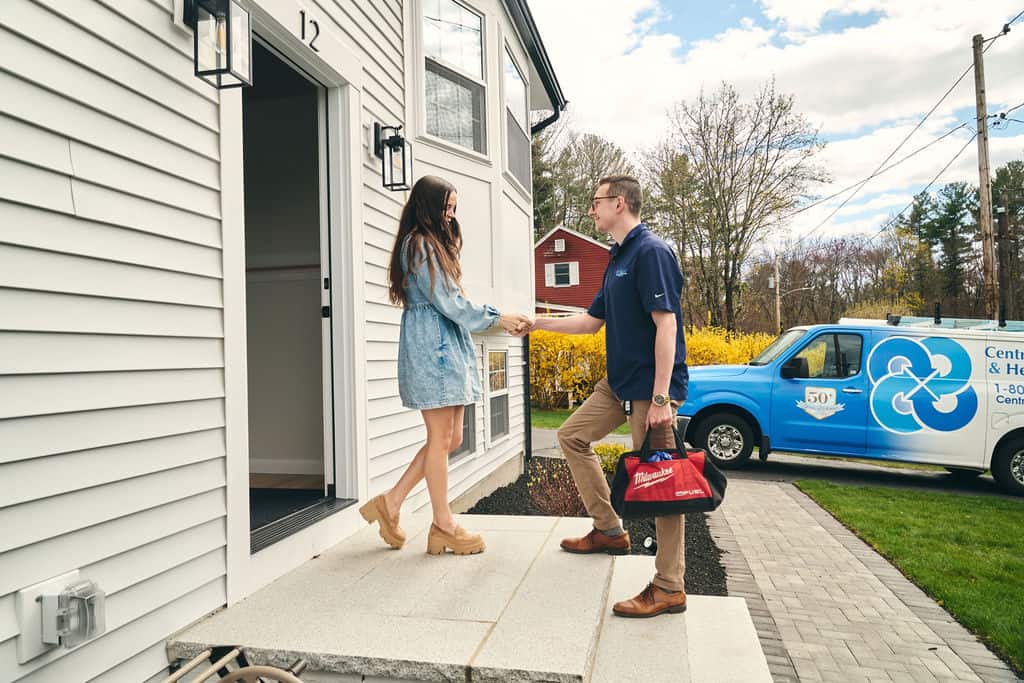 A woman greets a man delivering food at the door of a house, with a branded service van parked in the driveway.